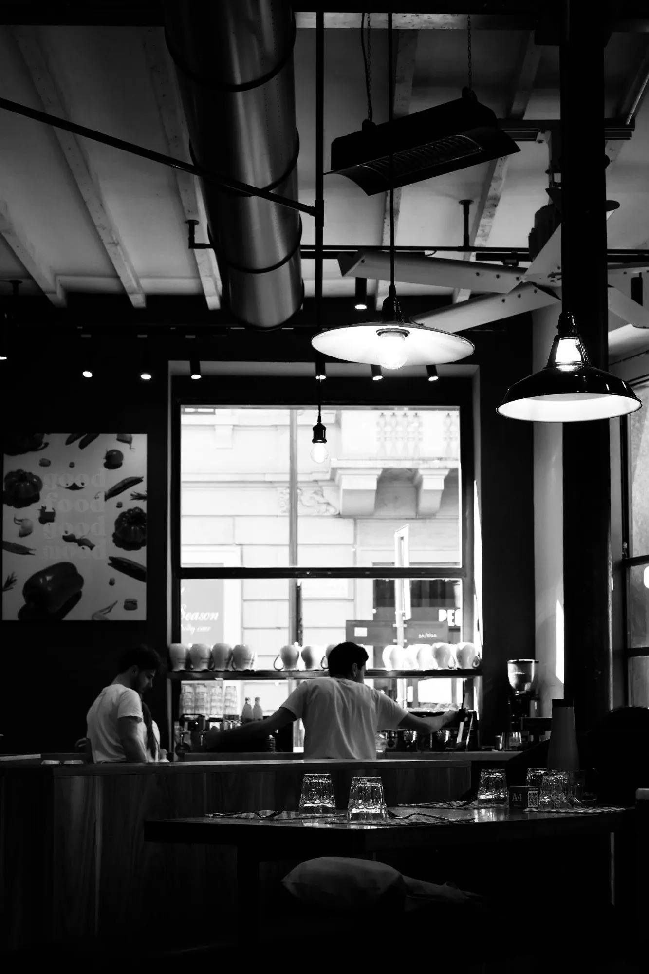 Two baristas working behind a cafe bar under pendant lamps, in black and white.